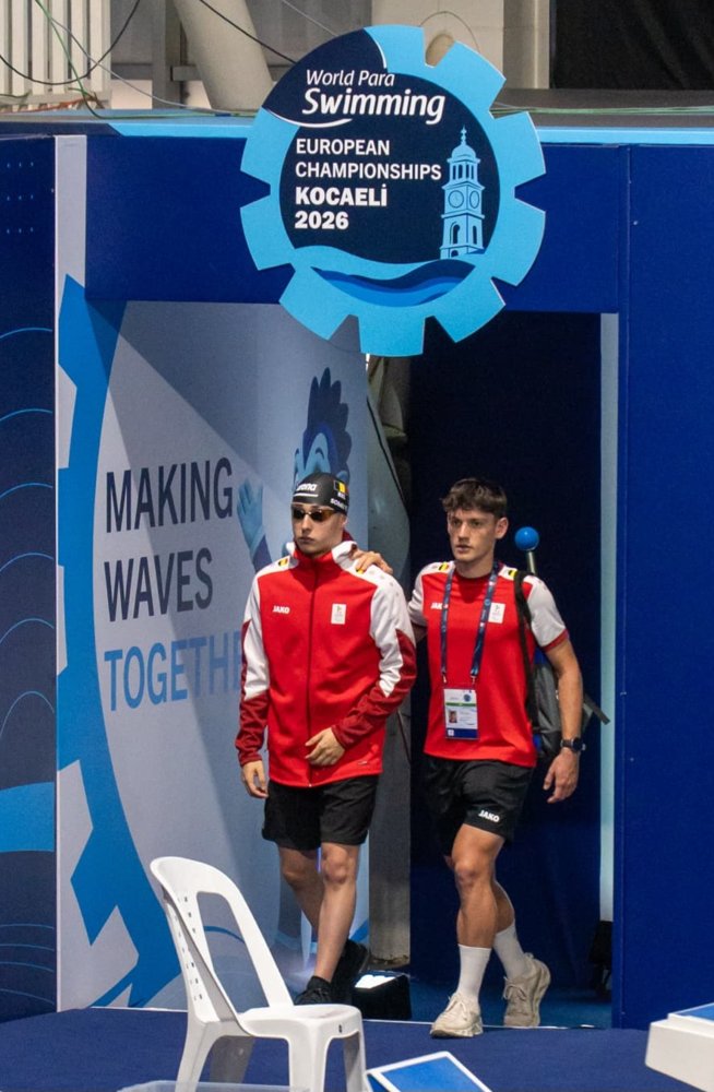 Thibaut Somei, met zwarte Belgische badmuts en zwembril, komt in het rode Team Belgium-trainingspak het zwembad van Kocaeli binnen. Zijn begeleider loopt naast hem met een hand op zijn schouder. Boven hen hangt het logo van de World Para Swimming European Championships Kocaeli 2026.
